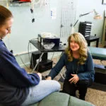 Two women conversing in a chiropractic clinic treatment room with therapy tables and medical equipment