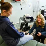 Two women in conversation inside a chiropractic clinic treatment room with medical equipment visible on walls and shelves