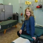 Woman in blue cardigan sitting on treatment table in chiropractic clinic with equipment on walls and another table visible in background