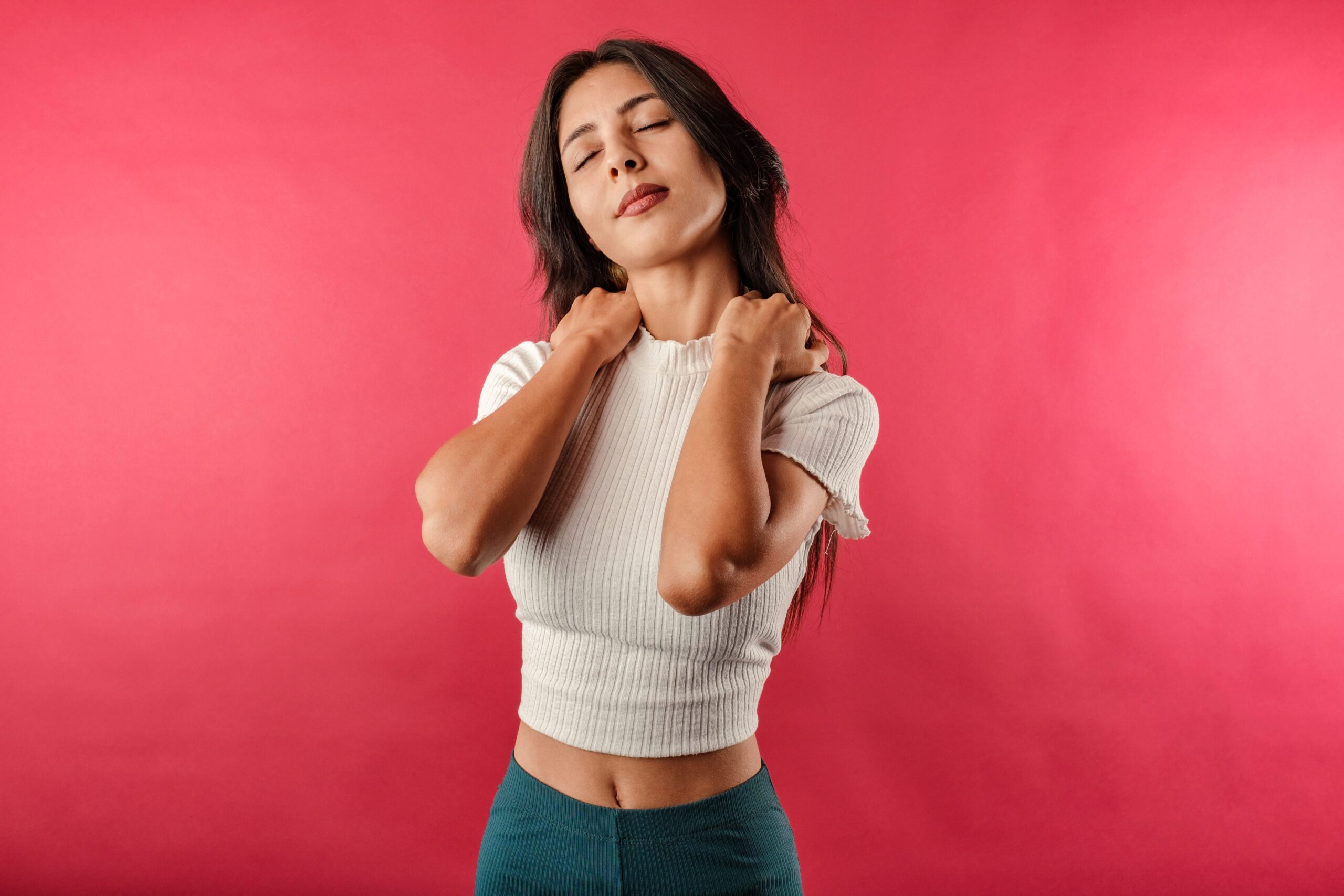 Young woman with eyes closed pressing fists against her neck and shoulders, showing signs of tension and muscle stress.